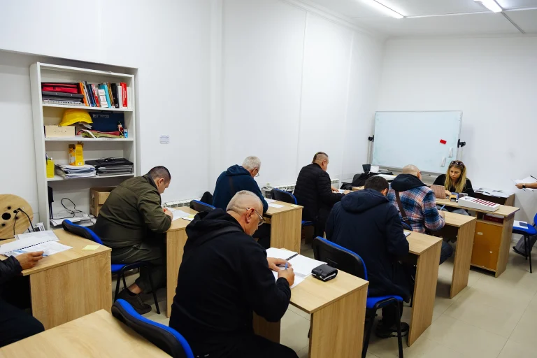 Класс с мужчинами и женщиной за партами, пишущими тесты, белая доска и полки с каской и книгами на стене
